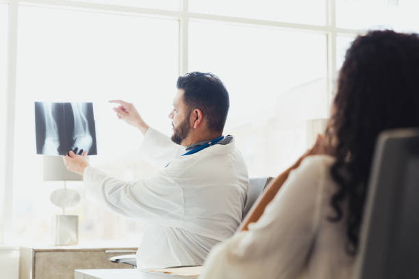 The mid adult male orthopedic doctor holds the patient's x-ray up to the light in order to show the unrecognizable female where the problem is.