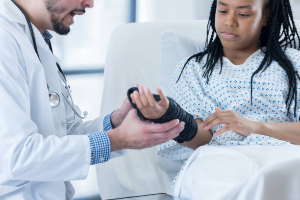 A serious teenage girl wears a hospital gown and lays in a hospital bed as she holds her arm out to her unrecognizable physician. Her doctor is showing her how to put on her new wrist brace.