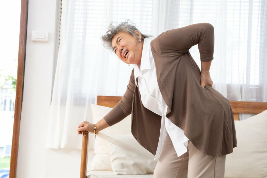 Image of a senior Asian woman bending over with her hand on the couch due to lower back pain, osteoarthritis, and spinal stenosis.