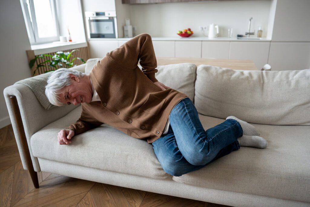 Elderly man struggles to rise from couch due to spinal stenosis pain.