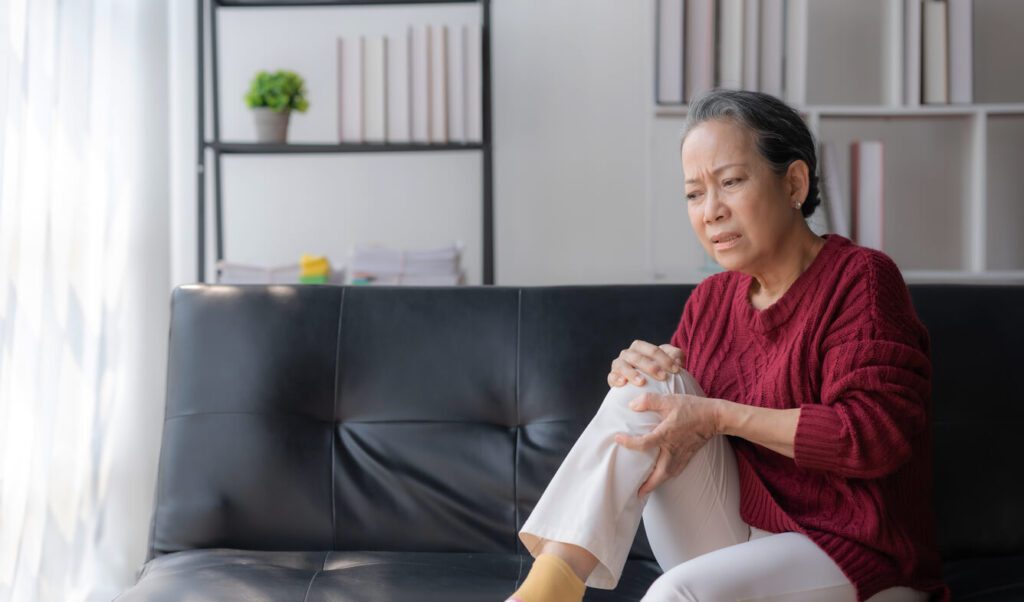 An older woman living with rheumatoid arthritis sits on a sofa at home, holding her knee in discomfort.