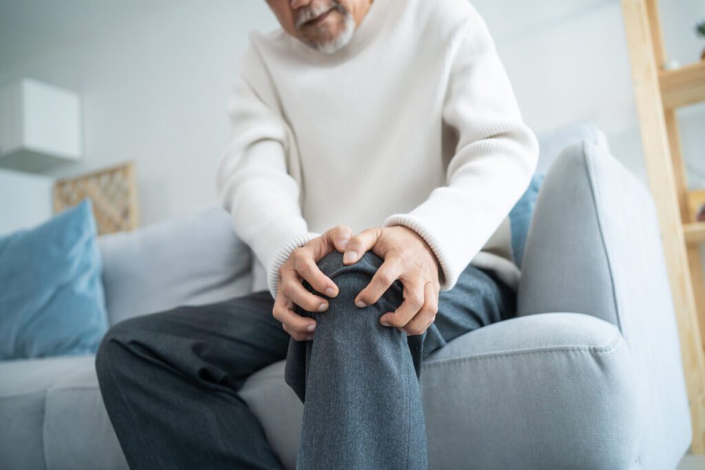 An elderly senior man with a disability sits on a sofa, experiencing knee arthritis.